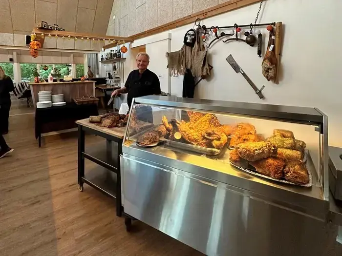 En kok står bag en montre fyldt med stegt kød i et rustikt rum, der er dekoreret med falske våben, pels og Halloween-tema.