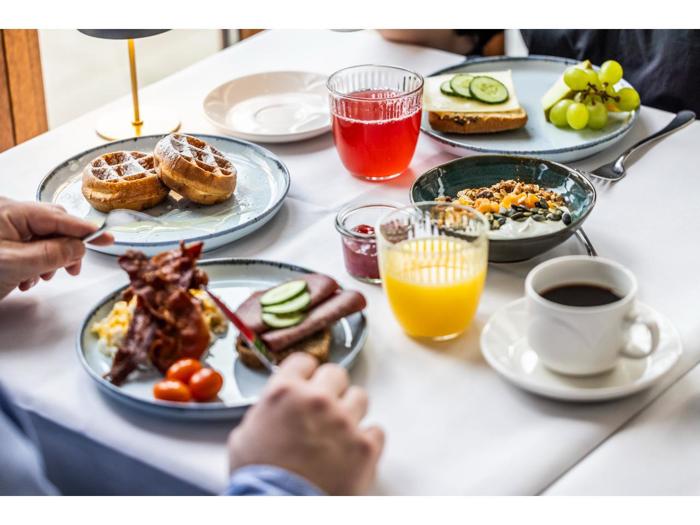 Et morgenbord med vafler, æg, bacon, tomater, toast med agurk, yoghurt med granola, kaffe, appelsinjuice, rød juice, vindruer og to siddende personer.