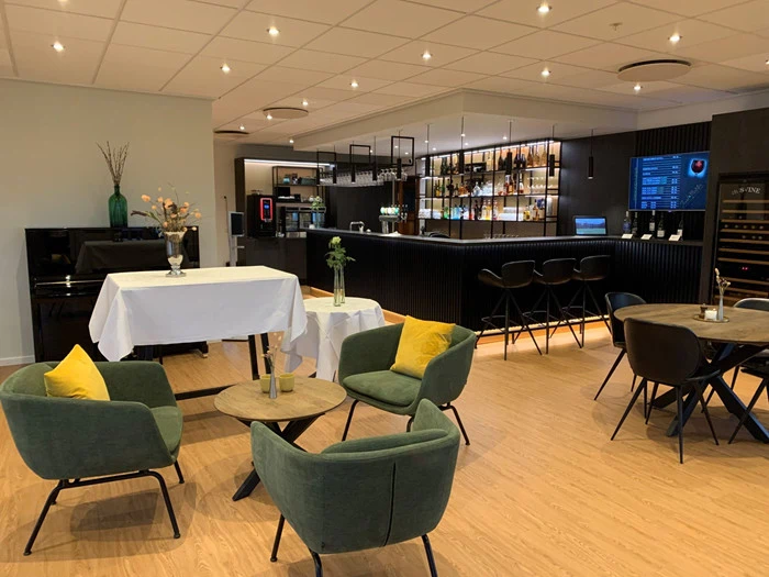 Moderne lounge med grønne lænestole, små runde borde og en bar i baggrunden. Rummet har trægulve, loftslamper og dekorative planter, som skaber en hyggelig og indbydende atmosfære.
