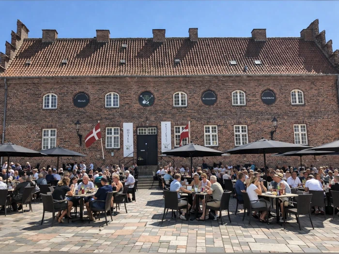Folk, der sidder ved udendørsborde foran et stort murstenshotel med det danske flag.