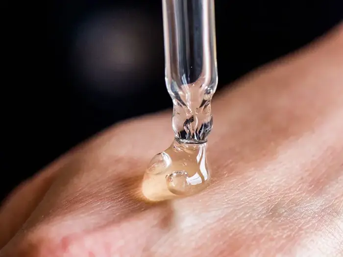 En dråbetæller, der doserer et klart serum eller en klar væske på bagsiden af en persons hånd, vist i nærbillede mod en mørk baggrund.