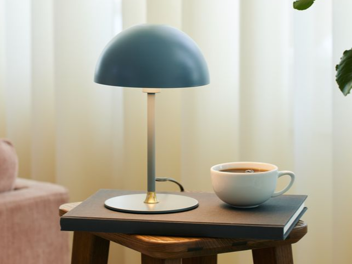 En moderne bordlampe og en kop kaffe hviler på en lukket bog, der er placeret på en træskammel ved siden af et svagt oplyst gardin.
