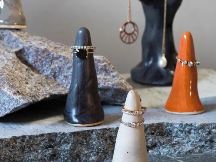 Tre keramiske ringholdere - sort, orange og hvid - viser ringe på en stenoverflade med smykker og flere holdere synlige i baggrunden.