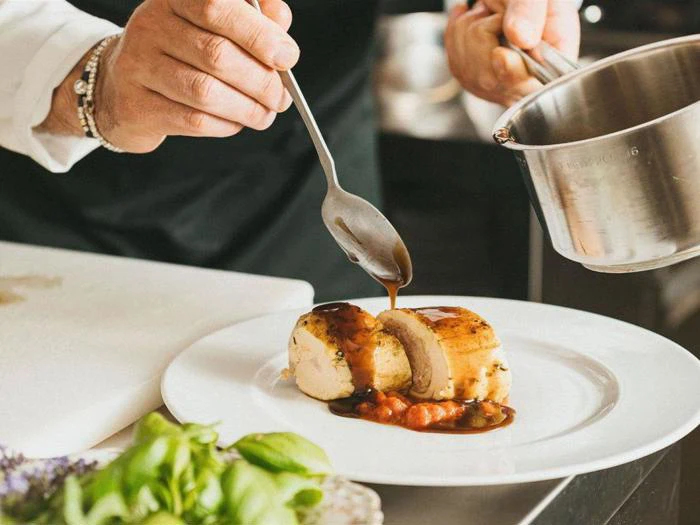 En kok hælder sauce over en fyldt kyllingeroulade på en hvid tallerken, pyntet med grøntsager, i et professionelt køkken.