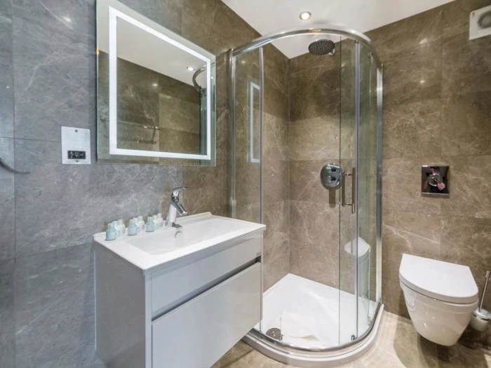 Moderne badeværelse med grå marmorvægge, brusekabine i glas, væghængt toilet og hvid håndvask med spejl over. Toiletartikler er pænt arrangeret på vasken.