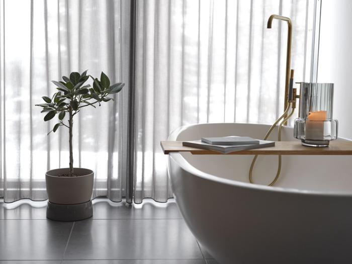 Moderne badeværelse med hvidt badekar, vandhane i guld, træbakke med bog og stearinlys, potteplante og gennemsigtige gardiner, der lukker naturligt lys ind.