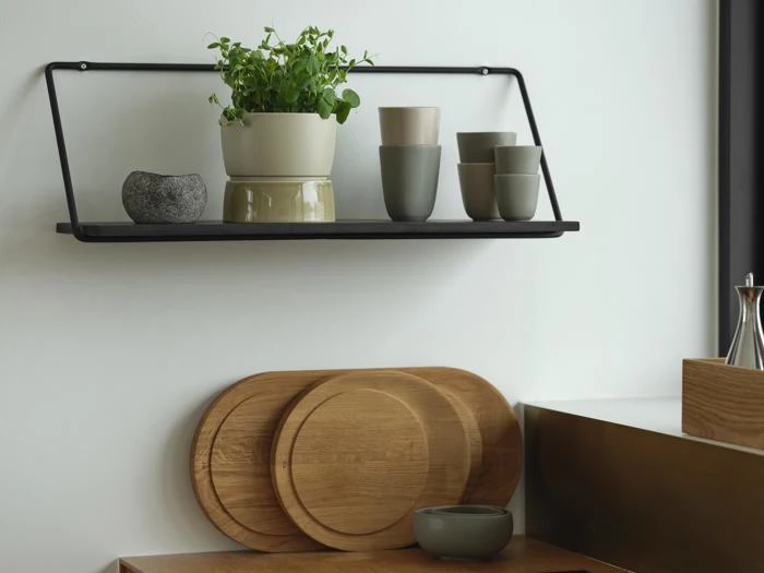 En sort væghylde rummer krydderurter i potter, kopper og en lille stenskål; nedenunder hviler træbakker og en skål mod en hvid køkkenvæg.