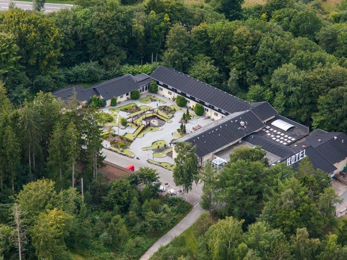 Luftfoto af en hotelbygning omgivet af tætte, grønne træer med anlagte haver og stier i gårdspladsen.