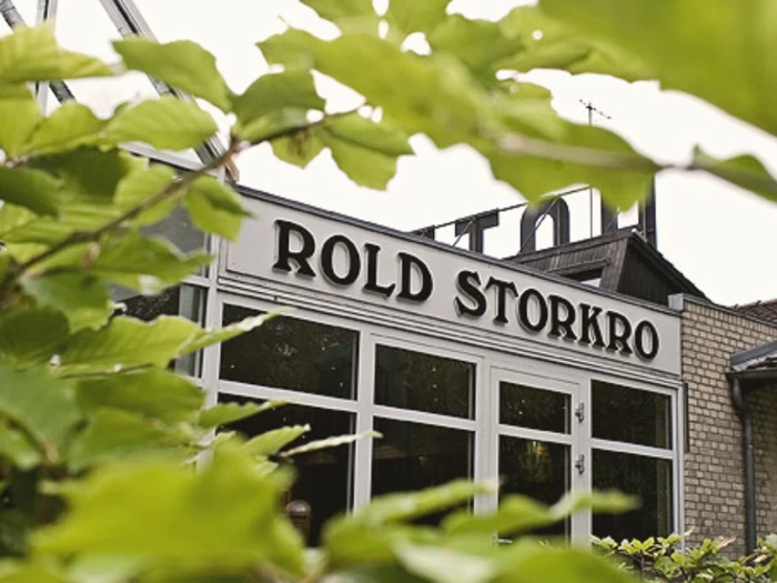 Ydersiden af en bygning med navnet Rold Storkro er delvist skjult af grønne blade i forgrunden.