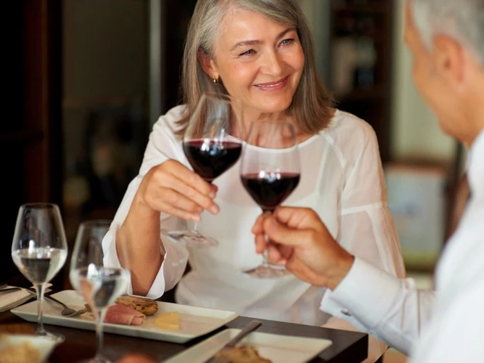 En ældre kvinde og en mand sidder ved et bord og smiler og skåler med rødvinsglas under et måltid.