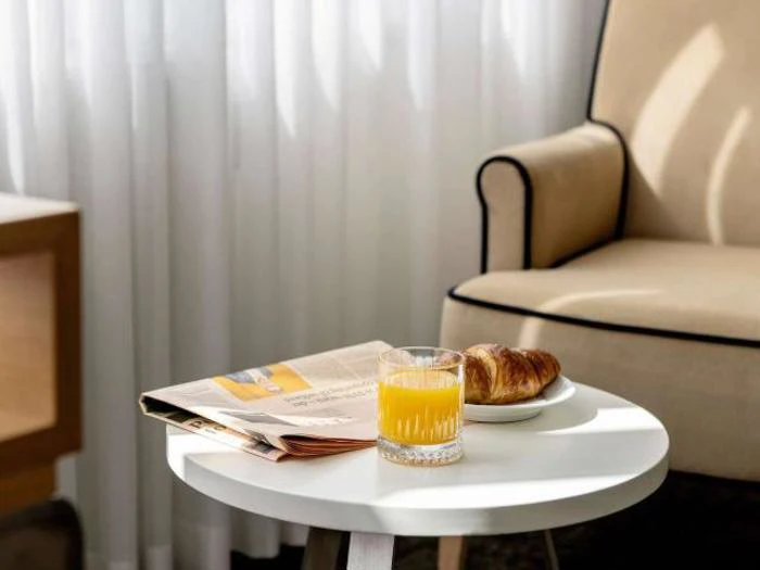 Et glas appelsinjuice, en croissant på en tallerken og en avis ligger på et lille hvidt bord ved siden af en beige lænestol med sort kant.