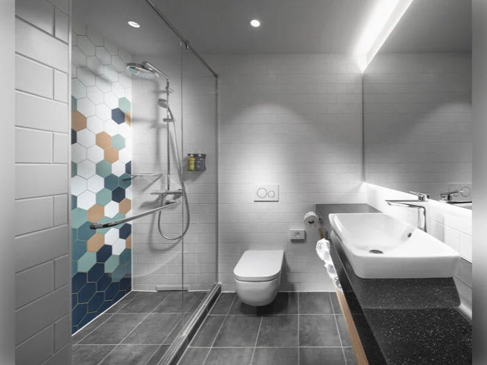 Moderne badeværelse med brusekabine med sekskantede fliser i blå, grå, brun og hvid, væghængt toilet, rektangulær vask og stort spejl over en sort bordplade.