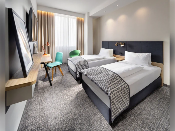 Moderne hotelværelse med to enkeltsenge, grå hovedgærder, hvidt sengetøj, mønstrede plaider, et skrivebord med to stole, et vindue med gardiner og indrammet kunst på væggen.