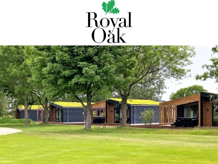 Moderne hytter med grønne tage er placeret blandt frodige træer og græs under et hvidt banner med Royal Oak-logoet og en grafik af egeblade.