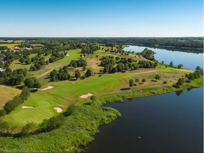 Luftfoto af en grøn golfbane med sandbunkers ved siden af en rolig flod, omgivet af træer og åbne marker under en klar blå himmel.
