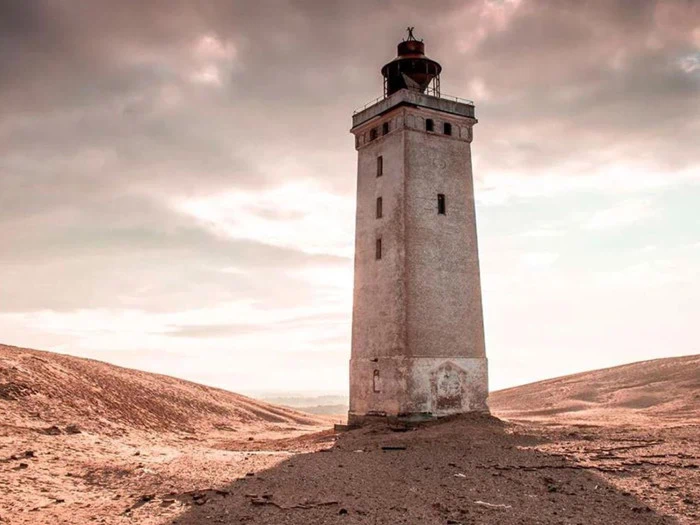 Et højt, forvitret fyrtårn står alene på sandet, gold jord under en overskyet himmel med blødt sollys.