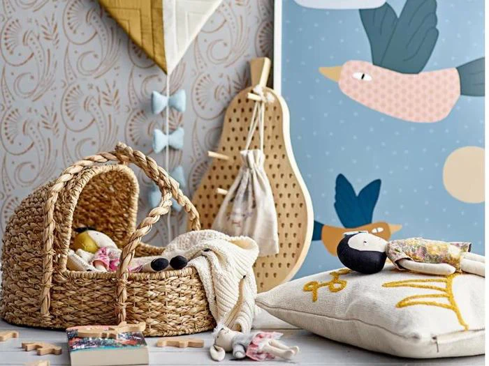 Et hyggeligt legeområde med en flettet kurv, et blødt tæppe, trælegetøj, en pude, en illustreret fugleplakat og dekorative vægophæng.