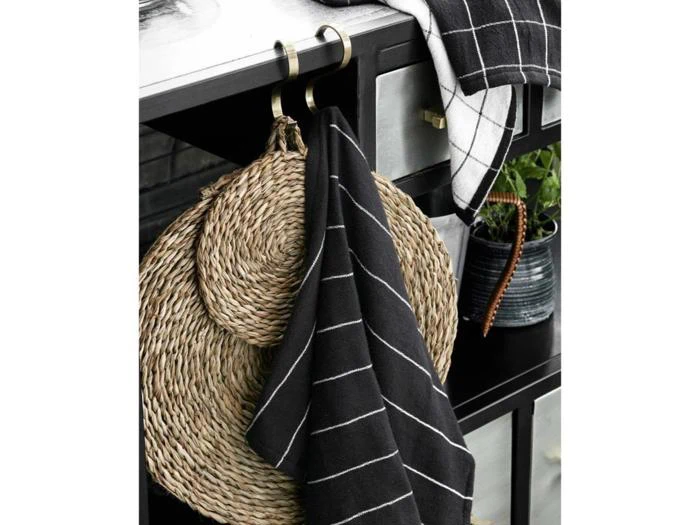 To runde, vævede dækkeservietter og et sort køkkenhåndklæde med hvide striber hænger på kroge fra et sort skab. På toppen af skabet står en potteplante og endnu et ternet håndklæde.