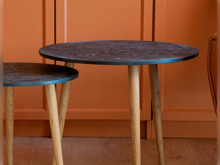 To runde borde med mørke plader og træben er placeret tæt på en væg med orange paneler og et trægulv.