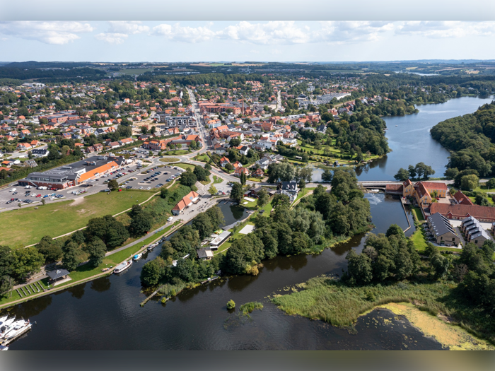 Luftfoto af en lille by med bygninger med røde tage, grønne træer, parkeringspladser og flere søer og floder, der snor sig gennem landskabet under en delvist overskyet himmel.