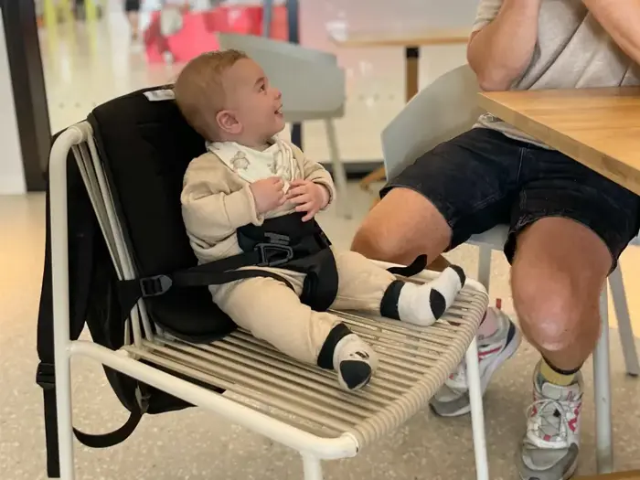 En smilende baby i beige tøj sidder sikkert i en sele på en stol og kigger op på en voksen, der sidder ved siden af dem ved et bord i et lyst indendørs miljø.