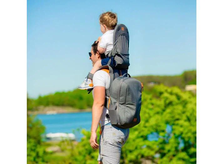 En mand bærer et lille barn på sine skuldre udendørs, begge iført afslappet tøj, med en rygsæk på ryggen og en naturskøn sø og træer i baggrunden.