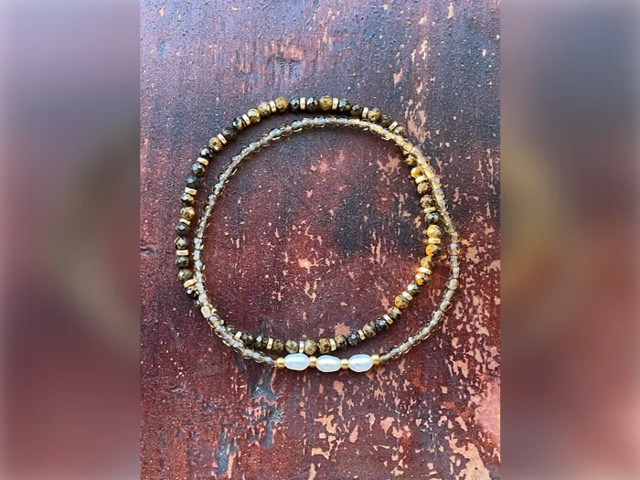 En perlekæde med små brune og guldfarvede perler og tre hvide ovale perler er arrangeret i en løs spiral på en rustik rødbrun overflade.