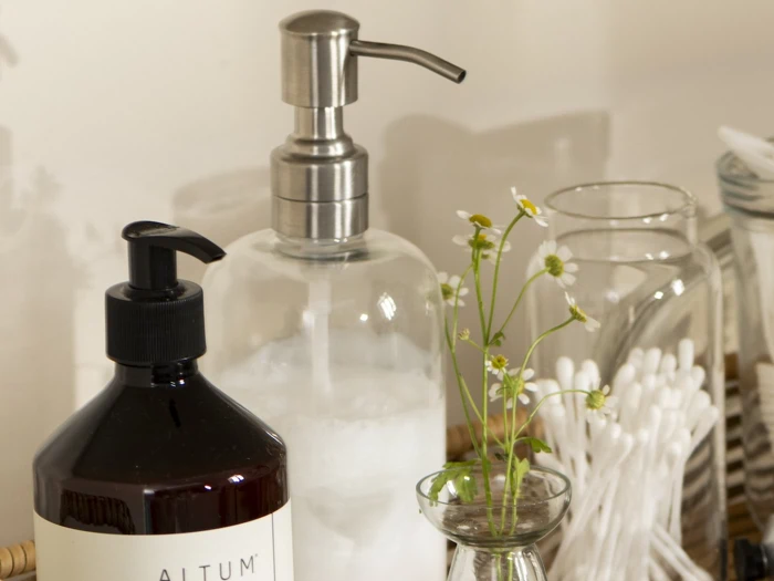 En sæbedispenser, en lotionflaske, en lille vase med kamilleblomster, glaskrukker og vatpinde arrangeret pænt på en bakke.