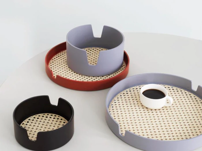 Tre runde bakker med vævede bunde i sort, rød og lilla, hvoraf den ene indeholder en hvid kop sort kaffe, er anbragt på en hvid flade.