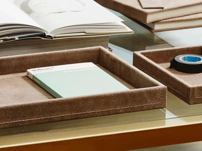 Nærbillede af et skrivebord med bakker i beige ruskind med en guidebog, sort tape og en åben bog på en glasplade.