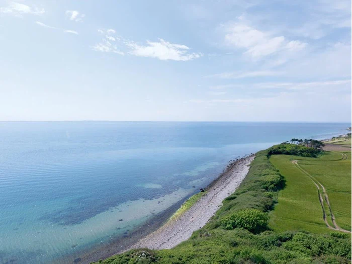 Et kystlandskab med klart blåt hav, en stenet kystlinje og frodige grønne marker under en delvis overskyet himmel.