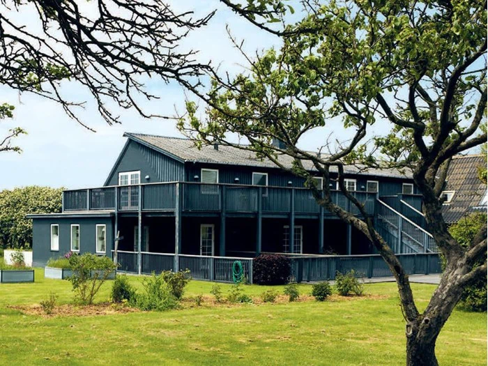 Et mørkeblåt træhus i to etager med altan, omgivet af grøn græsplæne, buske og løvtræer under en delvis overskyet himmel.