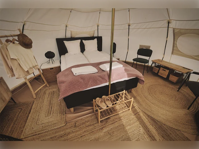 Et hyggeligt glampingtelt med dobbeltseng, lyserødt tæppe, håndklæder, stole, træopbevaring, vævede tæpper og pænt arrangeret tøj og tøfler.