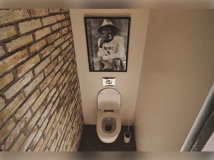 Et toilet med åbent låg er placeret under et indrammet sort-hvidt foto af en mand med hat, der er hængt op på en almindelig væg ved siden af fritlagt murværk. En toiletbørste står ved siden af toilett