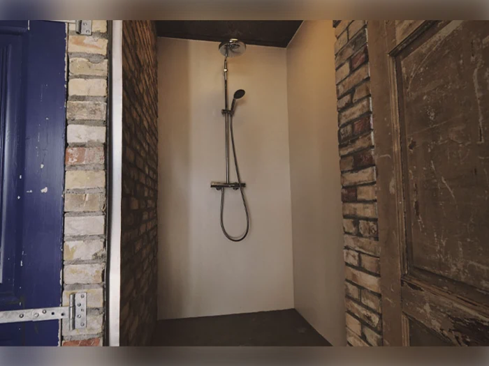 En enkel bruser med et håndholdt brusehoved i en lille alkove, omgivet af mursten og træbeklædte vægge og døre.