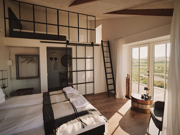 Et moderne, solrigt soveværelse med hems, store glasvinduer, trægulv, dobbeltseng og en stige, der fører op til øverste etage.