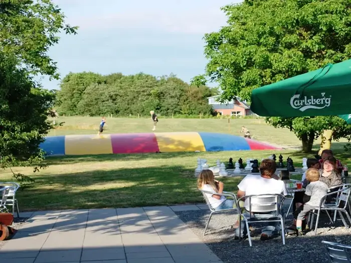 Folk sidder ved udendørs borde nær et gigantisk skakbræt, mens børn leger på en farverig oppustelig pude i en græsklædt park omgivet af træer.