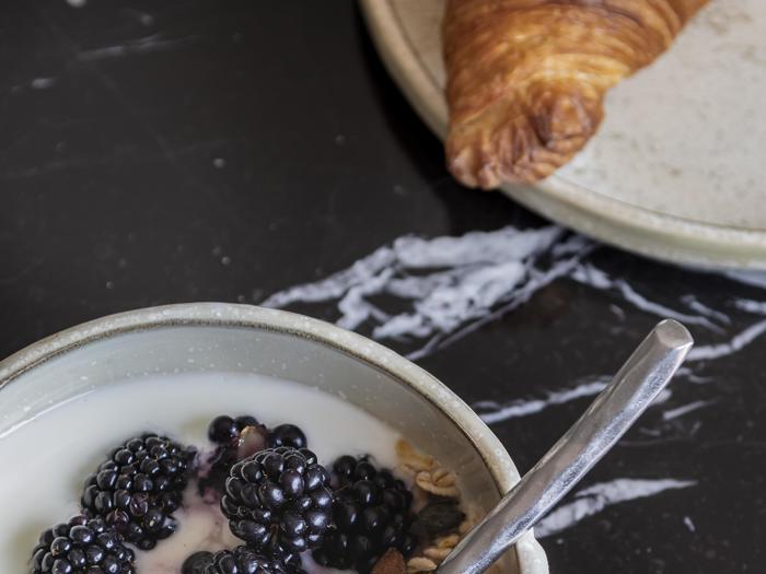 En skål cornflakes med mælk og brombær ved siden af en tallerken med en croissant på et mørkt marmorbord.