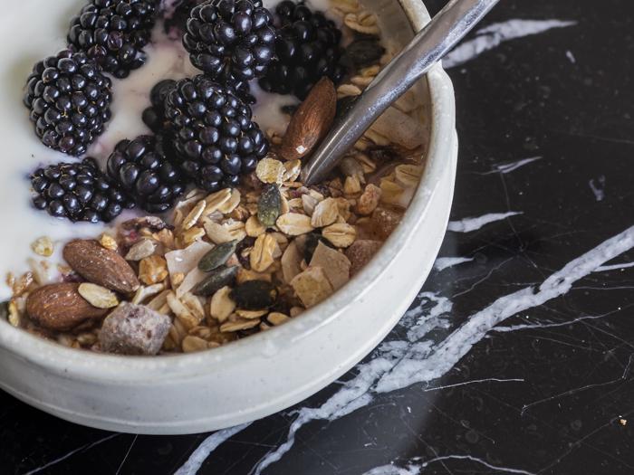 En skål med granola med mandler, frø og brombær toppet med mælk, placeret på en sort marmoroverflade med en ske indeni.