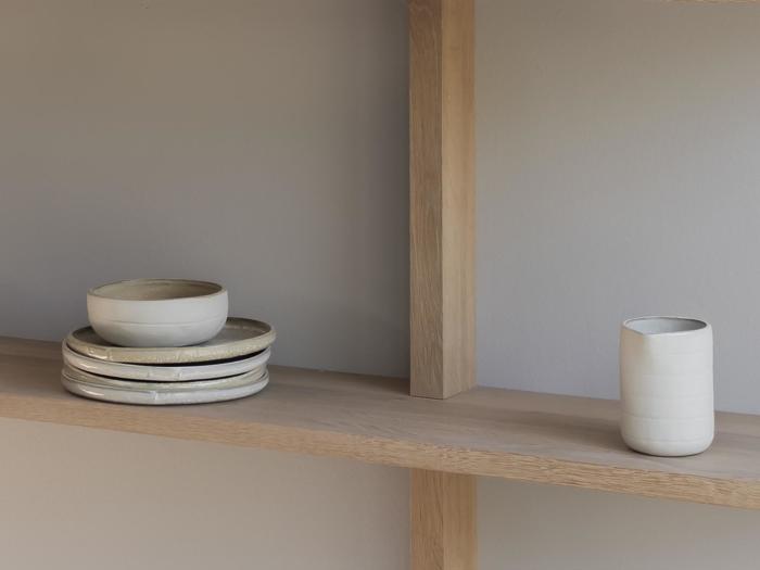 På en træhylde står en stak keramiktallerkener med en skål ovenpå, og en høj hvid kop står til højre.