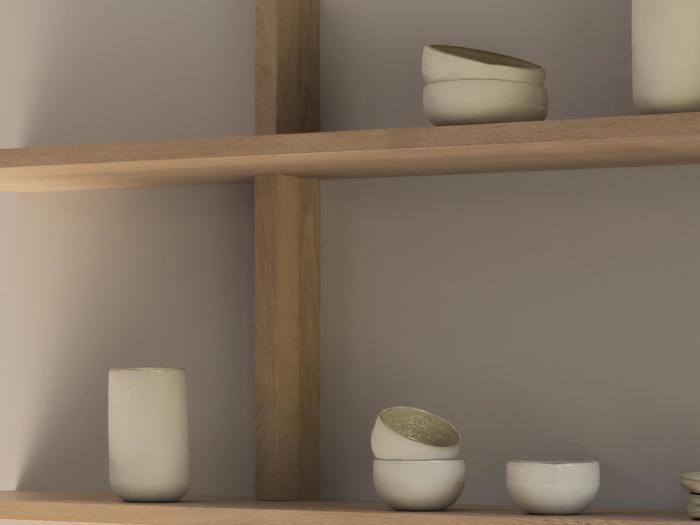 Minimalistiske træhylder med enkle, håndlavede hvide keramiske kopper og skåle, med dæmpet belysning og en neutral væg i baggrunden.