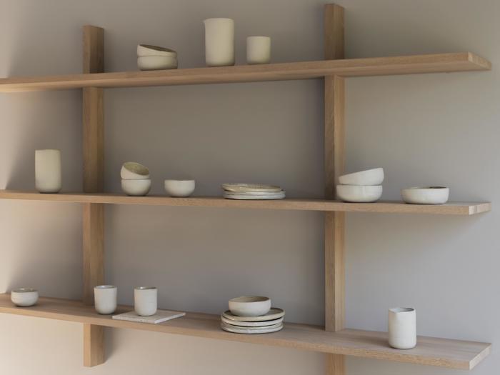 Tre træhylder viser minimalistiske hvide keramikvaser, skåle og kopper i forskellige former og størrelser mod en lysegrå væg.