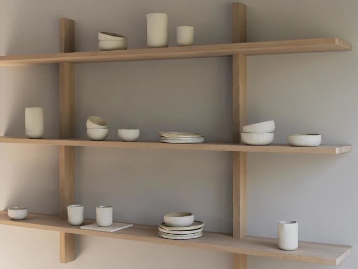 Tre træhylder viser minimalistiske hvide keramikvaser, skåle og kopper i forskellige former og størrelser mod en lysegrå væg.