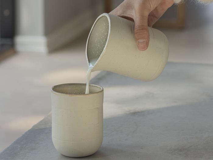 En hånd hælder mælk fra en keramikkande over i en matchende keramikkop på en lysegrå overflade.