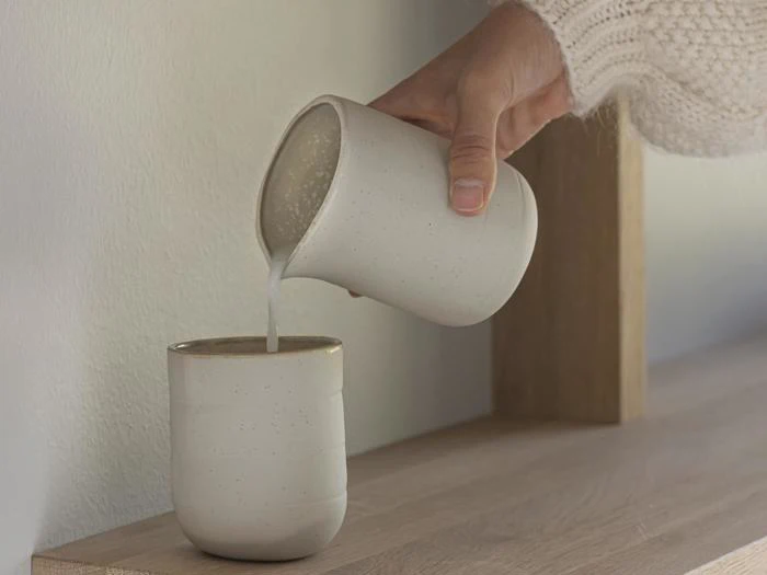 En hånd i en hvid sweater hælder mælk fra en keramikkop over i en anden keramikkop på en træplade.
