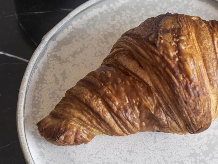 En gyldenbrun croissant ligger på en lys keramiktallerken med et spættet mønster oven på en mørk overflade.