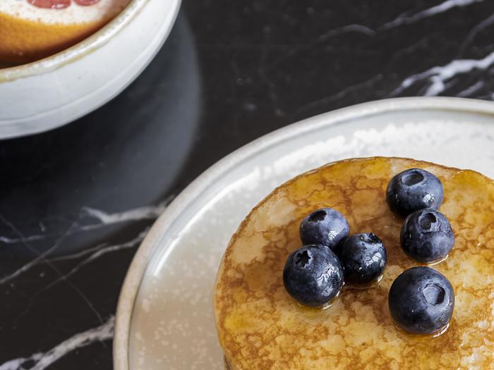 Nærbillede af pandekager med friske blåbær og sirup på en hvid tallerken med en skål grapefrugt i baggrunden på en sort marmoroverflade.