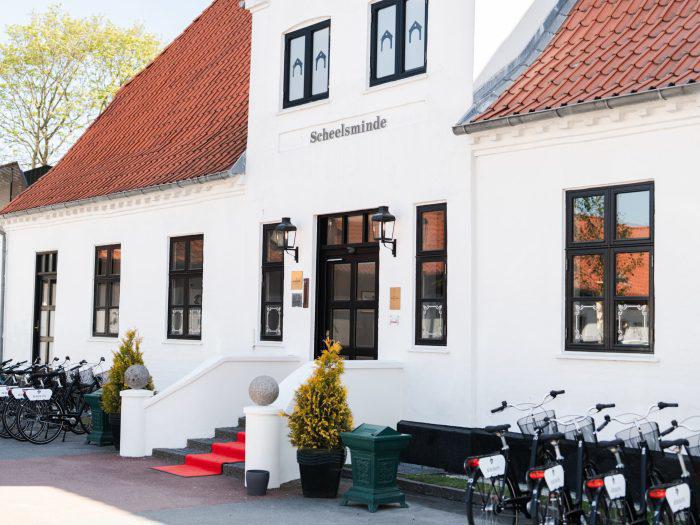 En hvid bygning med røde tagsten, mærket Scheelsminde, med et rødt tæppe på trappen, potteplanter og cykler parkeret langs siden.