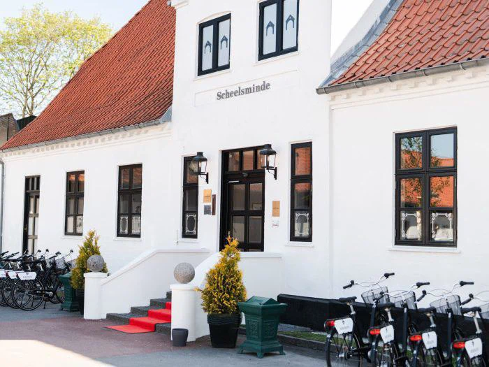 En hvid bygning med røde tagsten, mærket Scheelsminde, med et rødt tæppe på trappen, potteplanter og cykler parkeret langs siden.