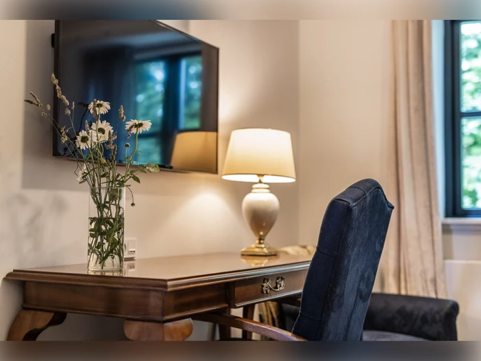 Et træskrivebord med en vase med blomster, en lampe og en stol står ved et vindue under et væghængt tv.